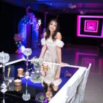 A woman elegantly poses in a formal setting with ambient lighting and decorations, indicating a celebration.