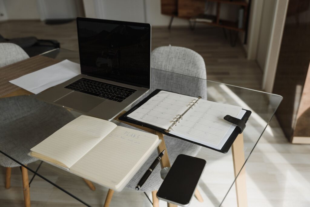 A clean and organized office setup with a laptop, notebook, and planner on a glass desk.